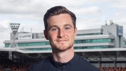 A headshot of Joe Jackson wearing a navy top with the Brentford Football Club logo patch on the the Umbro logo in white. He is in focus standing in front of a football pitch and stadium which is blurred in the background.